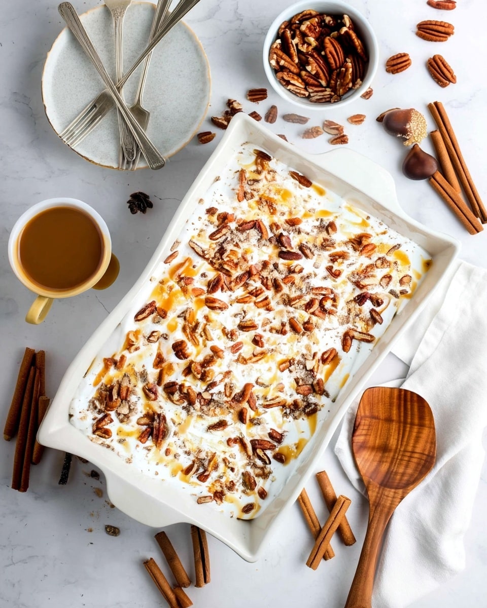 A rectangular white ceramic dish holds a dessert with three visible layers: a thick white creamy layer covers the entire surface, topped with scattered dark brown pecan halves and crushed nut pieces that add texture. Drizzled golden caramel sauce creates uneven streaks across the top, adding shine and contrast against the white cream. The dish is placed on a white marbled surface surrounded by cinnamon sticks, loose pecans, a wooden spatula, a cup of light brown coffee, and a white plate with silver forks. photo taken with an iphone --ar 4:5 --v 7