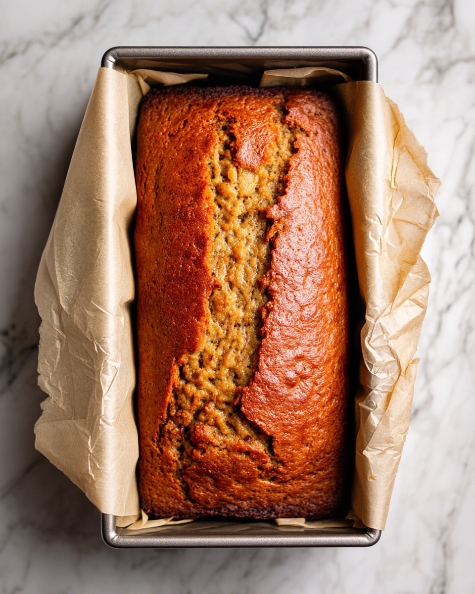 A freshly baked loaf of brown banana bread with a cracked top crust sits inside a parchment paper-lined metal baking pan. The bread's surface is textured, showing a golden to deep brown gradient from the sunken middle to the edges. The parchment paper is crinkled and light tan, folding over the sides of the pan. The background is a white marbled texture. photo taken with an iphone --ar 4:5 --v 7