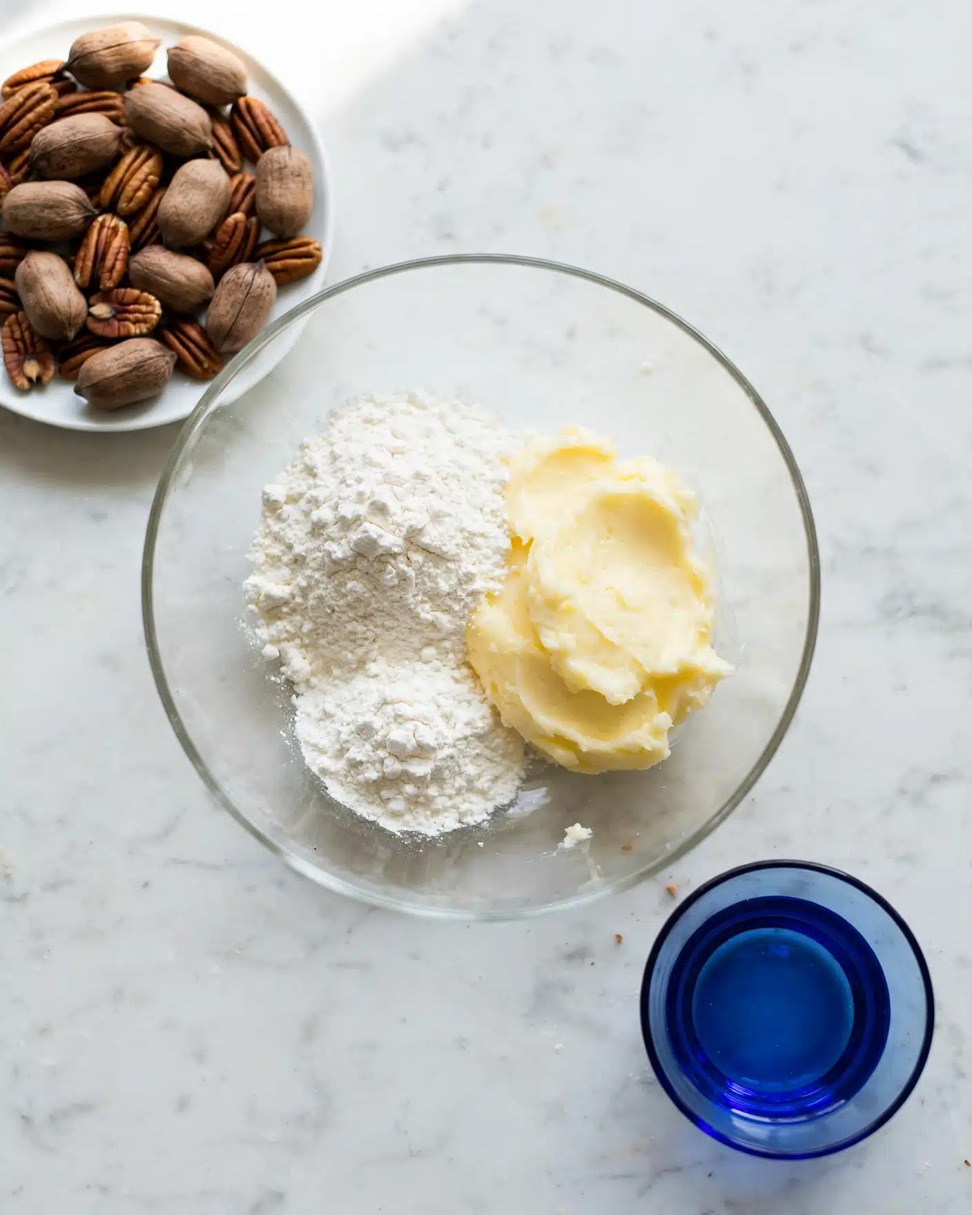 The image shows a clear glass bowl on a white marbled surface, containing three main layers: a pale yellow, soft butter-like layer on the right, a white powdery layer on the left, and another mound of white powder just below the first, all forming a small cluster inside the bowl. Near the bowl, to the top left, there is a white plate filled with whole pecans, showing their brown and textured shells. A small blue glass bowl is placed near the bottom right of the glass bowl, adding a touch of color to the scene. The overall setting is clean and minimalist, emphasizing the ingredients. Photo taken with an iphone --ar 4:5 --v 7