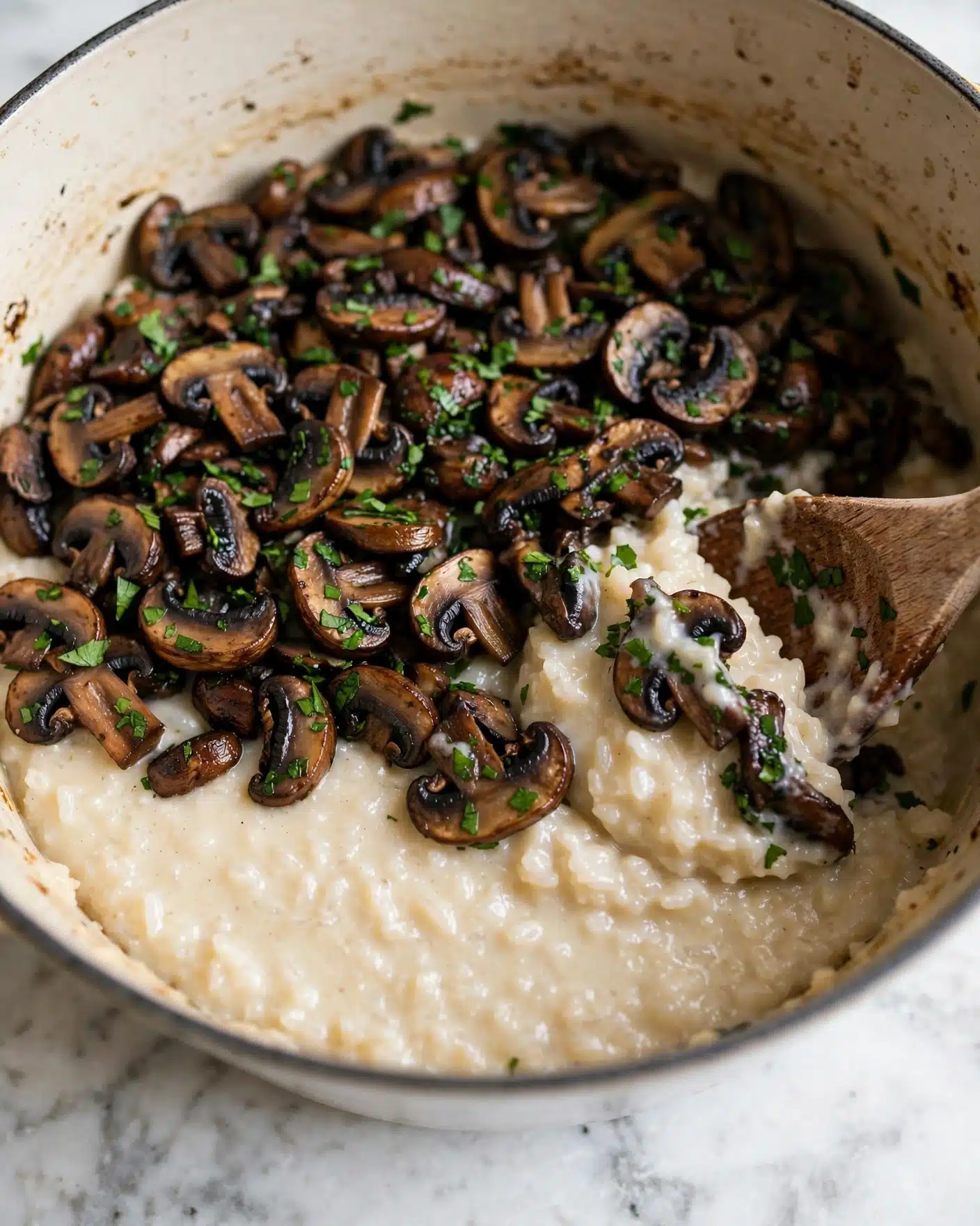 A close-up of a creamy dish in a pot showing two layers: a thick, light beige creamy rice base with a smooth texture and a top layer of sautéed dark brown mushrooms mixed with small bright green herb bits scattered evenly. A wooden spoon is partially dipped into the dish on the right side, slightly lifting some of the creamy rice and mushrooms. The inside of the pot is light beige with small marks from cooking. The whole scene sits on a white marbled surface. Photo taken with an iphone --ar 4:5 --v 7