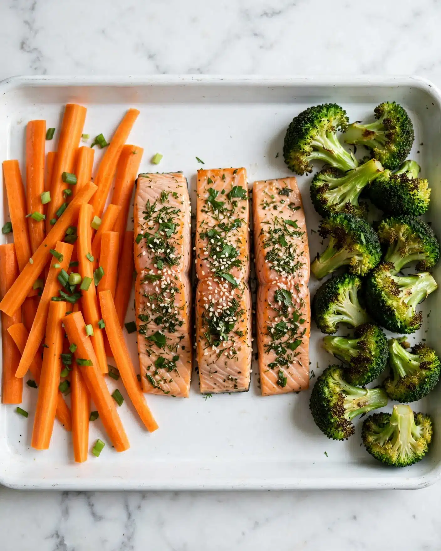 A baking tray filled with four salmon fillets in the middle, each pink with light grill marks and sprinkled with chopped green herbs and white sesame seeds. On the left side, there are thin, bright orange carrot sticks, also garnished with green herbs. On the right side, there are several bright green broccoli florets with slightly roasted edges. The whole tray sits on a white marbled surface. photo taken with an iphone --ar 4:5 --v 7