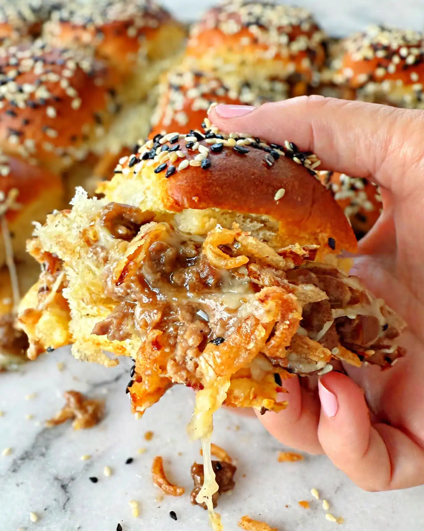 French Onion Sloppy Joe Sliders Recipe 5 The image shows golden brown rolls with a soft texture, topped with a mix of black and white sesame seeds. One roll is held by a woman's hand, revealing its inside layers, which include a crispy golden layer, melted cheese, and beef with a brown sauce. The rolls are placed on a white marbled surface with other similar rolls surrounding them in the background. Photo taken with an iphone --ar 4:5 --v 7