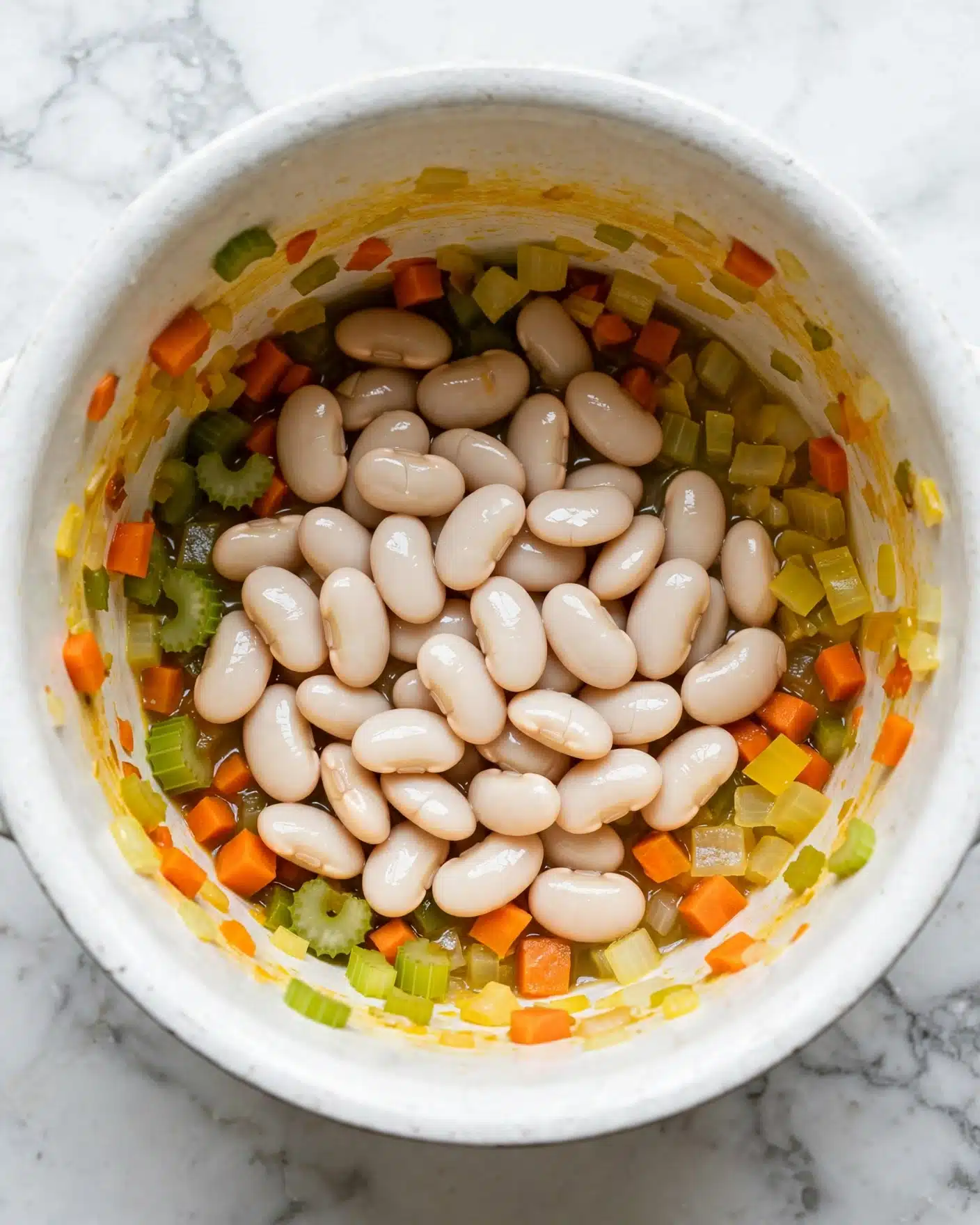 Inside a white pot on a white marbled surface, there is a mix of large white beans and small chopped vegetables. The vegetables include orange carrot pieces, green celery, and yellow onion bits, spread around the beans and sticking slightly to the pot's sides. The beans look smooth and shiny, sitting mostly in the middle with the colorful vegetables partly under and around them. The inside of the pot shows a yellow tint from the cooking process. Photo taken with an iphone --ar 4:5 --v 7