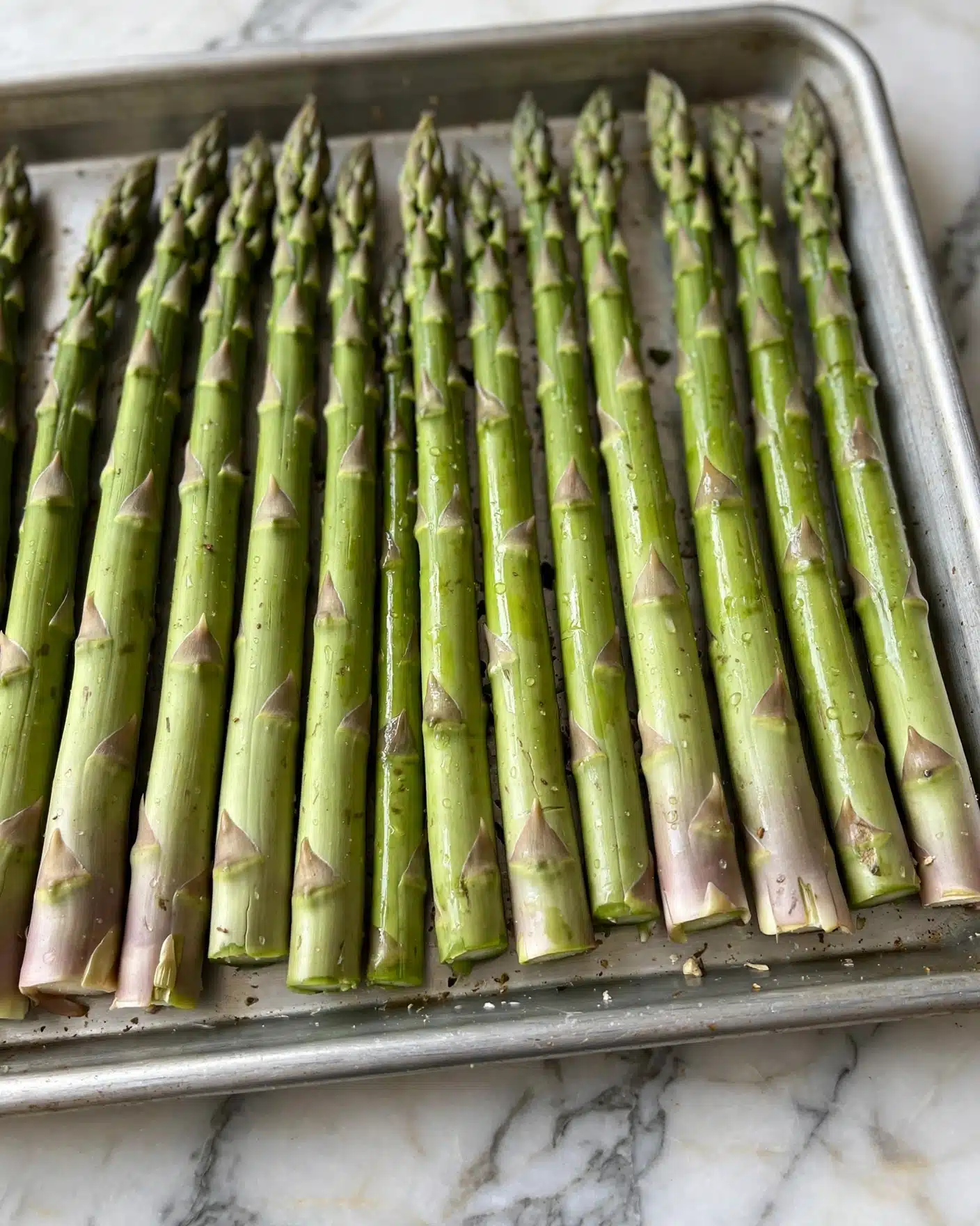A close-up image shows a bunch of raw green asparagus spears arranged side by side in a single layer on a baking tray, each spear featuring a textured, shiny surface with varying thickness and a pointed tip. The tray sits on a white marbled surface, highlighting the asparagus's vibrant green color and natural markings. Photo taken with an iphone --ar 4:5 --v 7