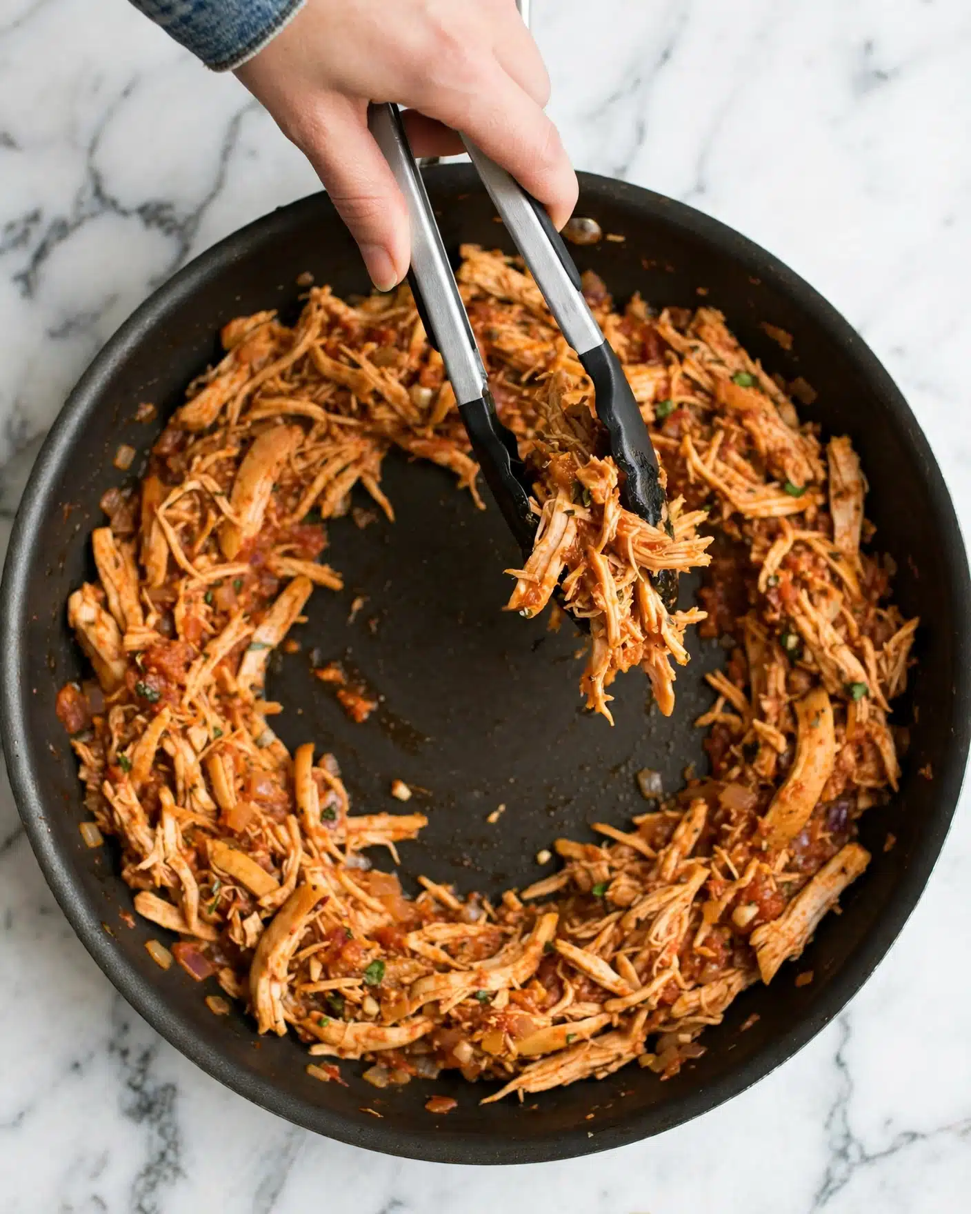 A dark non-stick pan is filled with shredded orange-brown cooked chicken, mixed evenly and spread mostly around the edges, leaving some space in the center. A woman's hand is holding silver tongs with black tips, lifting some of the chicken in the pan. The background is a white marbled surface. photo taken with an iphone --ar 4:5 --v 7