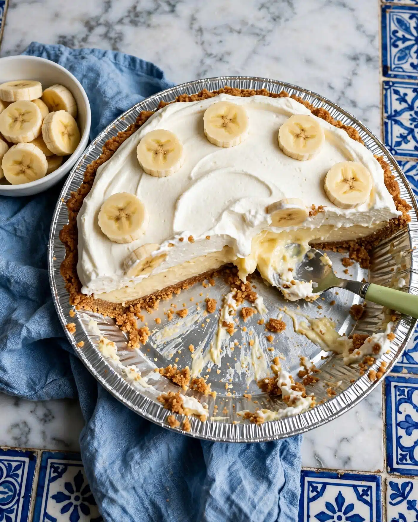 A pie in a round foil pan with three visible layers: a bottom light brown crumb crust, a thick creamy pale yellow filling in the middle, and a fluffy white topping spread unevenly on top. The top layer is decorated with round slices of banana scattered mainly around the edges. A section of the pie is missing, showing the layers clearly. The pan is placed on a dark blue cloth on a white marbled surface with a blue patterned cloth underneath. In the background, there is a white bowl with banana slices, a glass of milk, and a stack of white plates with two forks on top. Photo taken with an iphone --ar 4:5 --v 7