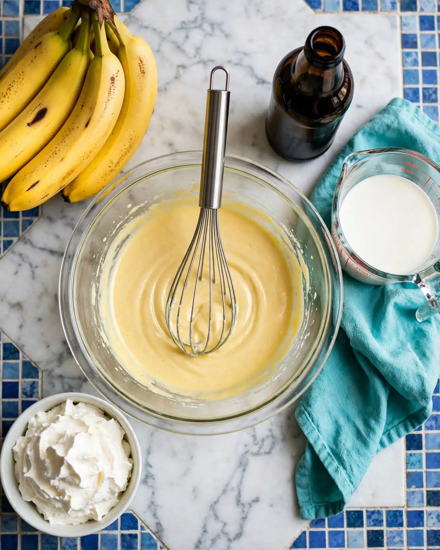 The image shows a clear glass mixing bowl in the center filled with a smooth yellow batter, with a metal whisk resting inside. To the left, there is another clear bowl filled with white fluffy cream. Above the bowls, a bunch of ripe yellow bananas is placed on a white marbled surface with a blue patterned cloth beneath the bowls. On the right side, there is a small dark bottle of vanilla extract with its cap off on the surface, next to a clear glass measuring cup containing some liquid. A teal cloth napkin is laid next to the measuring cup. Photo taken with an iphone --ar 4:5 --v 7
