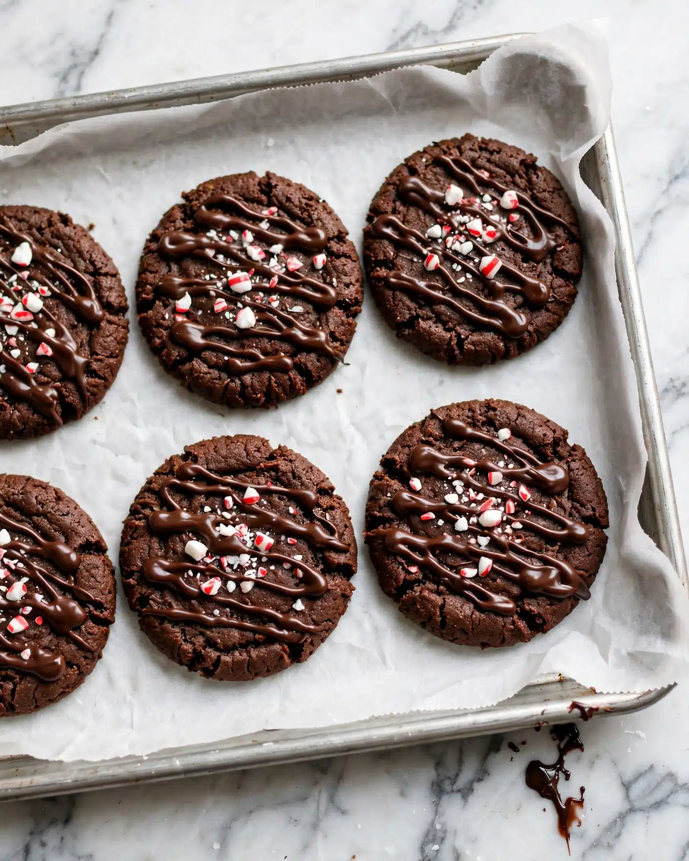 Triple Chocolate Candy Cane Cookies Recipe 4 There are six round chocolate cookies laid out on a baking tray lined with white parchment paper. Each cookie is dark brown and topped with a rough drizzle of dark chocolate, which creates shiny, wavy lines across the surface. Small pieces of crushed red and white peppermint candies are scattered on top of the cookies, adding pops of bright red and white color. The edges of the cookies are slightly raised and have a textured, crumbly look. The tray sits on a white marbled surface with a small drip of chocolate near the bottom right corner. Photo taken with an iphone --ar 4:5 --v 7