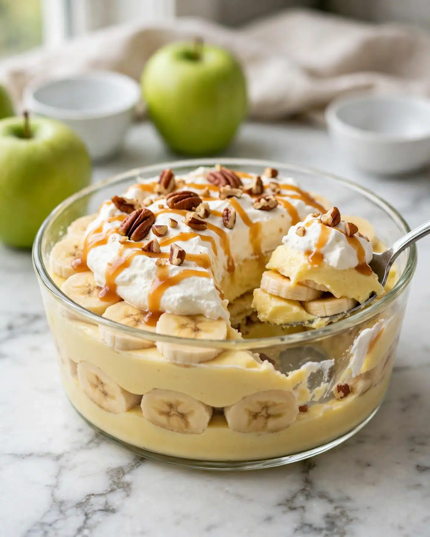 This image shows a clear glass dish filled with a layered dessert. The bottom layer is light yellow pudding. Above it are soft banana slices and more pudding, creating alternating layers. The top layer is white whipped cream with caramel sauce drizzled over it. Small pieces of brown pecans are sprinkled on top. A silver spoon is scooping some of the dessert from the side. The dish is placed on a white marbled surface with green apples and small white bowls in the background. The photo is taken with an iphone --ar 4:5 --v 7